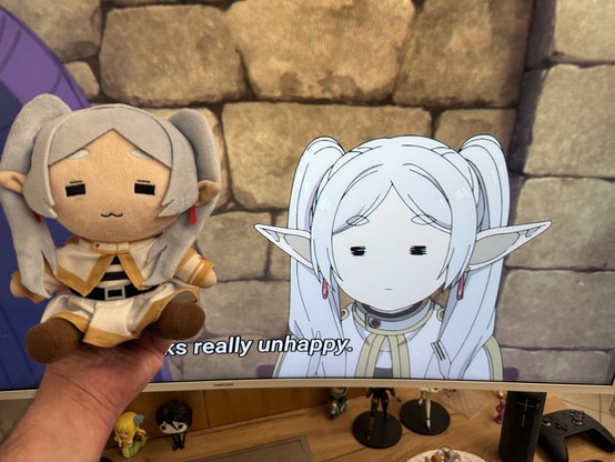 Picture of me holding the ยซย annoyed Frieren but happyย ยป plushie in front of a screen showing the ยซย annoyed Frierenย ยป scene from S02E02. I think the only difference is the mouth, W shaped for the plushie, small and flat for the anime :)
