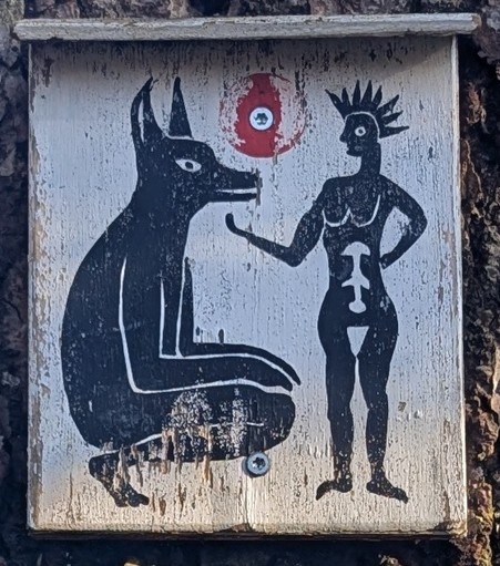 Das Bild zeigt eine kleine, rechteckige Holztafel, die an einem Baumstamm befestigt ist. Die Oberfläche ist weiß gestrichen, stark verwittert und weist Risse, Abplatzungen und Gebrauchsspuren auf.

Auf der Tafel sind zwei stilisierte, schwarz gemalte Figuren zu sehen:

Links sitzt ein großes, tierähnliches Wesen mit spitzen Ohren und länglicher Schnauze. Es erinnert an einen Hund, Schakal oder eine mythologische Figur. Der Körper ist kompakt, die Vorderbeine sind angewinkelt, und das Tier blick…