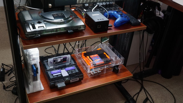 Two lighted shelves showing a Sega Mega Drive/Genesis (Model 1) in a smoked acrylic shell, a MiSTer FPGA, and a SCART switch with five SCART inputs and one SCART output being used.  The second shelf houses a PS1 (in a frosted acrylic shell), a SNES (in a smoked acrylic shell), and an NES (in a clear acrylic shell with a Ninten-drawer installed).