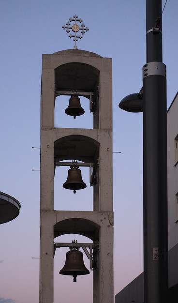 Ein Turm aus Betonsäulen mit einem Kreuz oben drauf. Der Turm hat drei Kammern, in jeder dieser Kammern eine Glocke, nach oben hin werden die Glocken kleiner.