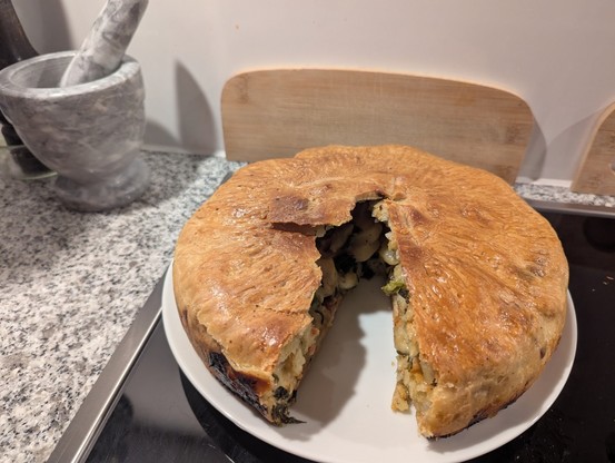 Auf dem Bild ist eine Cholera, ein traditionelles, rundes Ofengericht, das goldbraun gebacken auf einem weißen Teller liegt. Ein großes Tortenstück wurde herausgeschnitten, wodurch man die Schichten im Inneren gut erkennen kann. Die Füllung besteht aus Kartoffeln, Lauch und Äpfeln, die dicht und gleichmäßig in einem Teigmantel eingeschlossen sind.

Die Oberfläche ist knusprig und leicht glänzend, mit einer rustikalen, handgeformten Struktur. Am unteren Rand zeigt sich eine etwas dunkler gebacke…