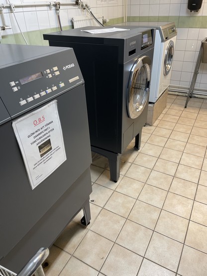 A laundry room featuring three modern washing machines in gray and white. The machines are side by side on a tiled floor, with a notice posted on the left machine. The walls are tiled with a green accent.
