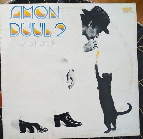 Foto des Plattencovers: Chris Karrer im weißen Anzug, der mit dem weißen Hintergrund verschmilzt. Er trägt schwarze Stiefeletten mit hohen Absätzen und einen schwarzen Hut. Dabei beugt er sich vornüber und lässt sich von einer auf den Hinterbeinen stehenden, schwarzen Katze, die ein überdimensioniertes und brennendes Streichholz in der linken Vorderpfote hält, die Zigarre anzünden.
Links oben in der Ecke Bandname und Albumtitel in einem blauen Font, der an Art Deco erinnert.