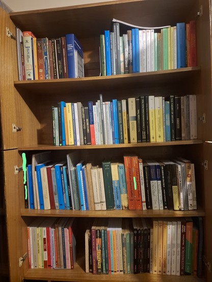 Vemos uma estante com quatro prateleiras quase completamente preenchidas com livros. Se percebe que a grande maioria deles é de matemática superior, mas vemos também um número considerável de livros de matemática de ensino médio e também alguns de programação e de ensino. Os livros estão dispostos de pé, facilitando que sejam colocados ou retirados. Também se percebe que houve um cuidado na organização destas prateleiras, com livros de áreas afins geralmente próximos entre si. Notamos também qu…