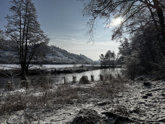 Sehr gerne 😊

Das Foto zeigt eine ruhige Winterlandschaft an der Vils bei Traidendorf. Im Vordergrund liegt das leicht verschneite Ufer mit gefrorenem Gras, Zweigen und unregelmäßigen Spuren im Schnee. Die Vils zieht sich ruhig und dunkel schimmernd durch das Bild, teils von dünnem Eis überzogen, und spiegelt das helle Winterlicht.

Links und rechts rahmen hohe, kahle Bäume die Szene ein. Ihre mit Reif überzogenen Äste glitzern in der Sonne und geben dem Bild eine fast märchenhafte Stimmung. Im…