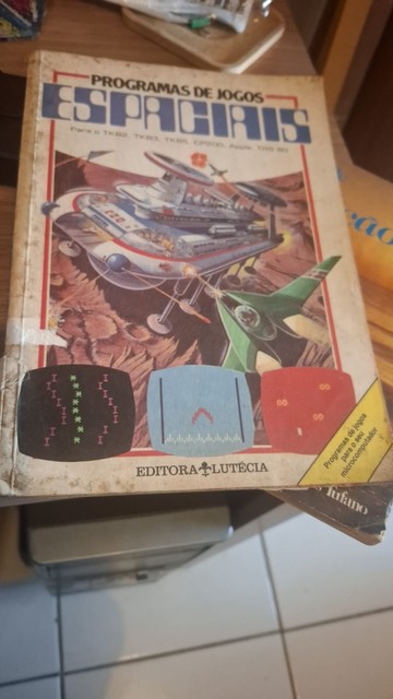Em cima de uma mesa vemos um livro com aparência de antigo e maltratado, cuja capa é de uma batalha de naves espaciais (em estilo bem retrô, que lembra até sci-fi dos anos 70) e três miniaturas sugerindo jogos especiais (também em estilo muito retrô). O título do livro é "Programas de jogos espaciais", com a observação abaixo de que são programas para os computadores TK82, TK85, CP200, Apple e TRS 80.