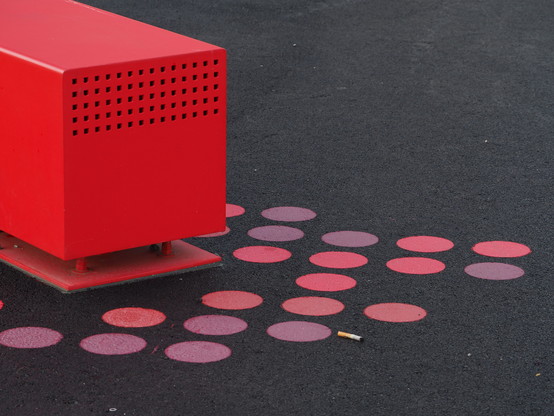 Ein roter Würfel im linken oberen Teil, rote Punkte auf dem dunkelgrauen Boden aufgeklebt, daneben ein Zigaretten-Stummel. 