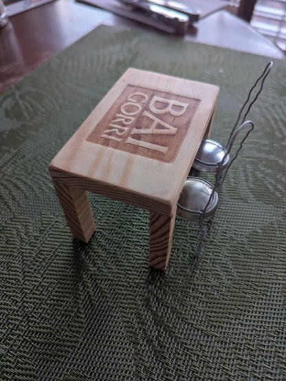 Ein kleiner Tisch aus dem hellbraunen, gemaserten Holz einer Weinkiste. Auf der Tischplatte ist die Inschrift "BAI GORRI" eingebrannt. Auf der linken Seite des Tischs sind sind zwei kleine Stühle untergeschoben, die aus dem Metallkäfig von Champagnerkorken gebastelt sind. Die beiden Stühle sind aus silberfarbigem Metall.
