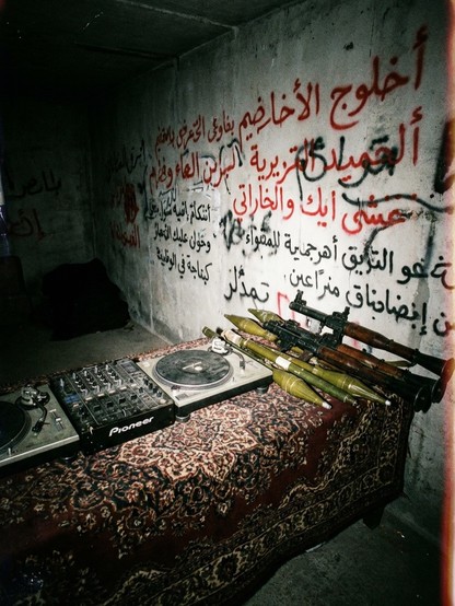 dark concrete nook with arabic graffiti on the walls, a dj setup on a traditional carpet draped over a table, and some rocket launchers