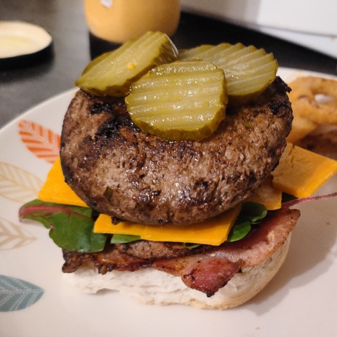 A beef burger in a bun sat on top of bacon, salad and cheese, with gherkins slices on top. 