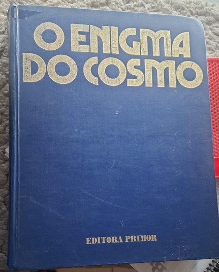 Um grande livro em capa dura e grossa, com o título de "O enigma do cosmo", da Editora Primor.
