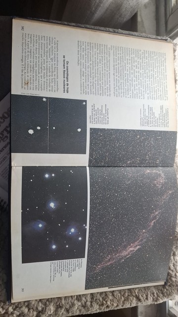 Um livro está aberto em uma página com astrofotografias coloridas. Uma delas é um close bem de perto das plêiades, que são belas com seu azul difuso característico de estrelas gigantes azuis. Há um pouco de texto, com um destaque em letras maiores dizendo que os cosmólogos de hoje se tornavam físicos nucleares.