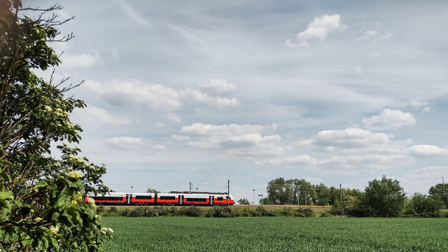 Landschaftsbild mit einem Zug im unteren Teil, oben viel Himmel, unten ein schmaler Streifen eines Getreidefeldes. 