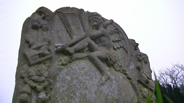 a gravestone. the carved words have weathered away but there remains a carved reaper with his scythe, reading from a book, an hour glass carved on either side.