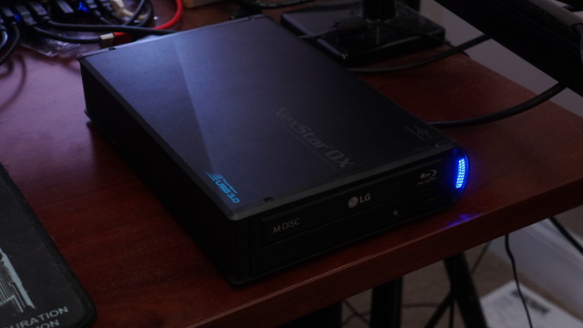 An LG WH16NS60 optical drive in an NexStar DX 5.25" USB3 enclosure sitting on top of a desk.  A violently bright blue LED flashes showing disc reading activity.
