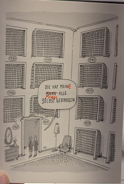 Eine schwarz-weiß Zeichnung, die einen Innenraum zeigt, mit Fussballtoren an den Wänden, aufgehängt wie Trophäen, wobei jede mit einem Jahr beschriftet ist: 1969, 1970, 1973, 1979, 1983, 1986, 1989 und 1990. Zwei Personen stehen in einem Durchgang unten links, neben einer Kommode mit einem Spiegel und einer Vase. Eine Stehlampe und ein Sessel befinden sich rechts. In einer Sprechblase in der Mitte steht der deutsche Text "DIE HAT MEIN MANN ALLE SELBST GESCHOSSEN", wobei "Mein" mit einem rot gef…