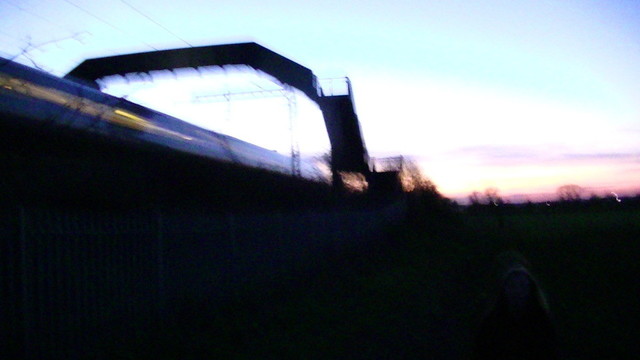 a blurry purple, orange and blue dusk with train speeding under a foot bridge. you can see the interior lights on the train as one long blurry line. every thing is like a dark silloette. idk how to spell silloette. i am tired now.