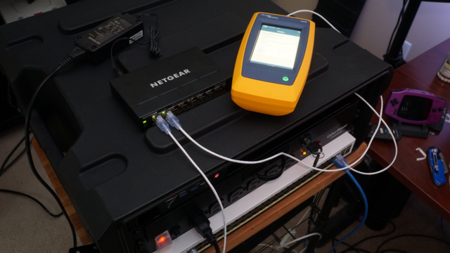 A picture of a Fluke network tester connected to the Netgear PoE switch showing a successful test.  The Netgear switch is connected to the main Cisco VoIP switch.