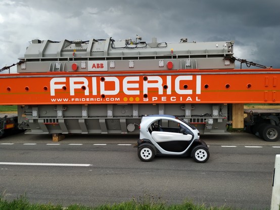Ein kleines, grauschwarzes Elektroauto Renault Twizy vor einem Spezaltransport eines riesigen Transformators. Der graue Transformator ist in einem orangeroten Rahmen eingebaut. Er ist 1.5 mal höher und etwa drei mal länger als das Auto.