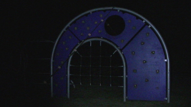 an 'n' shaped climbing frame in the dead of night, containing a wooden purple arch shaped climbing wall. the mouth of the arch has a rope climbing net stretched over it. and there is a big circular hole in the wooden arch. i suppose it looks a bit like an eye. goodnight.