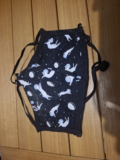Máscara facial de pano. A estampa é bonitinha e algo infantil, apresentando gatinhos brancos usando capacete de astronauta sob um fundo preto de espaço sideral, estrelas e afins.