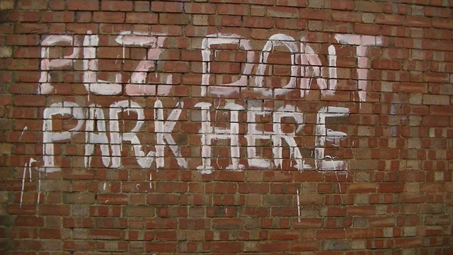 a red brick wall in some narrow victorian back road with the words "PLZ DONT PARK HERE" painted in a roughly mixed pink and white paint.