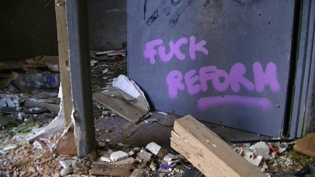purple "FUCK REFORM" graffiti at the bottom of a grey door in a condemned building full of crap