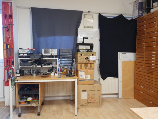 The picture shows Update's electronics corner. On the left side there is a small table with soldering and lab equipment. Next to the table, a few boxes are stacked with a 3D printer looking out of the top one. On the right wall there are wooden component drawers. On the wall behind the lab table there are blankets covering the windows. Amazingly, barely anything is lying around on the floor and only four boxes are visible in the picture.