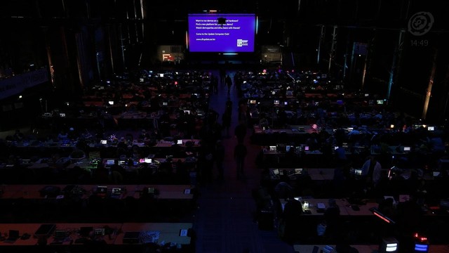 A screenshot of the Revision 2026 demoparty stream. The screenshot shows a big dark hall filled with rows of tables. People with computers are sitting at the tables. The only light in the pictures comes from the many computer screens and the big screen in the front of the hall. The big screen  shows a slide with the following content:

"Want to run demos on real oldskool hardware?
Find a new platform for your next demo?
Watch demoparties and drink beers with friends?

Come to the Update Compute…