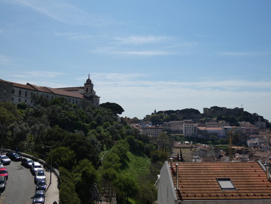 Una vista del castillo de Lisboa tomada desde una colina cercana. Se ve también una iglesia, tejados y árboles bajo un cielo azul