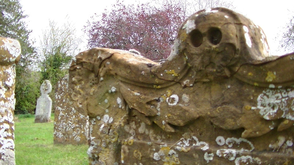 a grave stone topped with a carving of a skull, but the bottom half of the skull has fallen away. patches of white lichen. more graves in background.