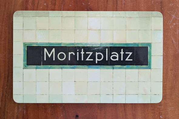 Foto eines Brettchens mit einem Motiv aus dem Berliner U-Bahnhof Moritzplatz. Der Schriftzug 'Moritzplatz' in weissen Lettern auf schwarzem Grund, darum ein grüner Rand. Um diese Kartusche herum die typischen hellgrünen Wandfliesen der BVG Bahnhöfe.
