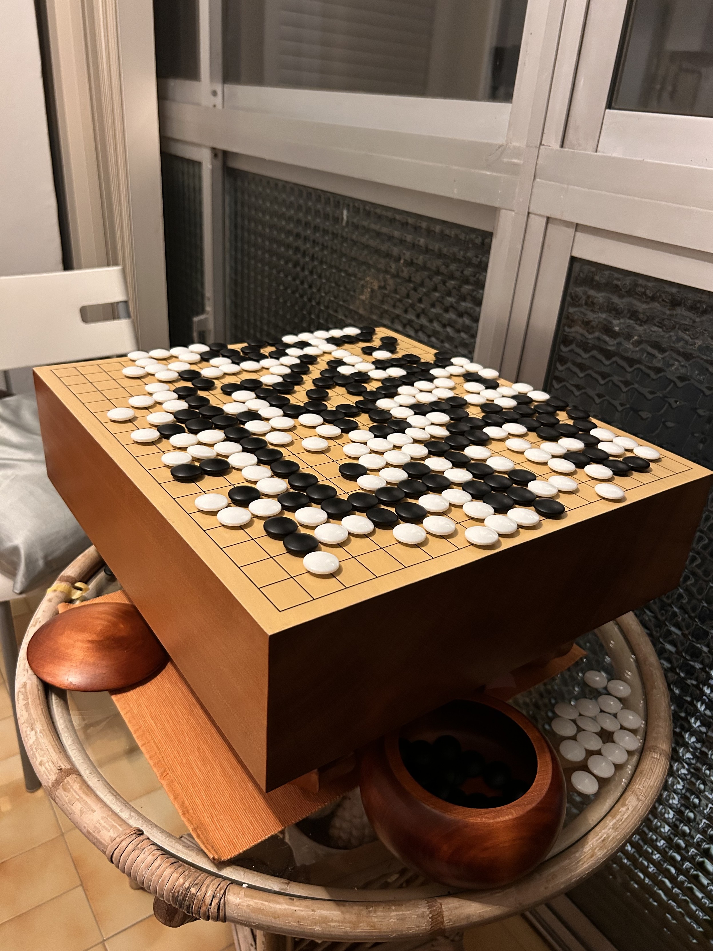 A thick wooden goban mid-game, covered in black and white stones in a complex position. Traditional wooden bowls sit beneath the board on a rattan table, photographed indoors at night.