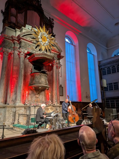 Foto fra koncerten: de tre udøvende musikker på scenen under prædikkestolen. En trommrslager, en bassist og en ved klaveret. Desuden ses nogle høje kirkevinduer med blå lys i. Nogle røde spotlights danner et rødt skær langs væggene og søjlerne.