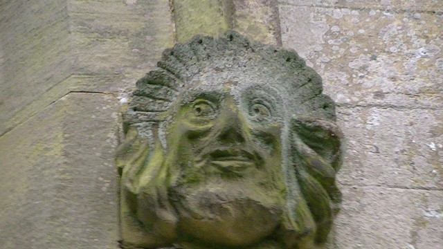 a stone carving of a daft looking face. it is next to a window on a church (out of shot)