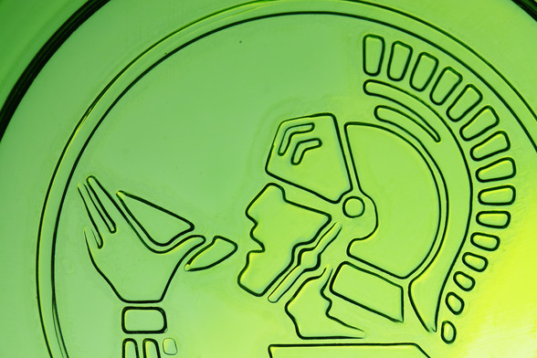 Eine grüne Fläche mit einer schwarzen Zeichnung darauf von einer Person, die aus einer Schale trinkt. Die Person trägt einen römischen Helm. Das Bild ist die Aufnahme vom Boden eines grünen Wasserglases. Der Römer ist das Logo einer Firma für Mineralwasser. 