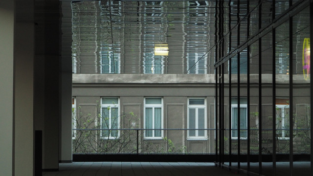 Ein "Durchblick" in einem Gebäude auf die Fensterfront des Nachbarhauses, wie in einem Rahmen. Die Fenster spiegeln sich auch noch in der Decke des "Rahmens". Die Farben sind beinahe monoton grau, nur die weißen Fensterrahmen bringen etwas Kontrast ins Bild. 