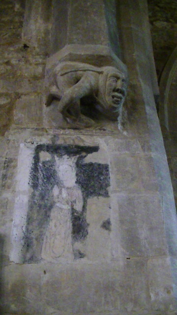 a grotesque medieval corbel. looks like a crouched monk or something with an oversized head, maybe tongue sticking out. below the corbel there is a medieval wall painting of a crowned lady. more like a ghost painting now.