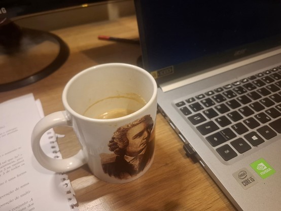 Vemos em cima de uma mesa parte de um notebook à direita da imagem e alguns papéis à esquerda. O foco da fotografia é em uma caneca branca decorada com uma imagem de Euler.