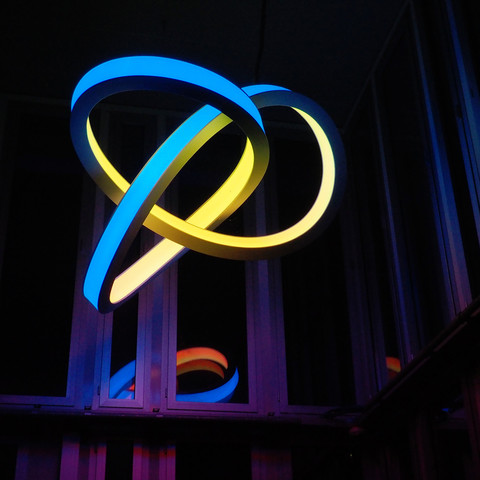 Eine Skulptur, die als Lampe dient. Es ist ein verschlungenes endloses Band, auf einer Seite gelb, auf der anderen blau.  Sehr dunkler Hintergrund, die Aufnahme wurde abends bei Dunkelheit gemacht.