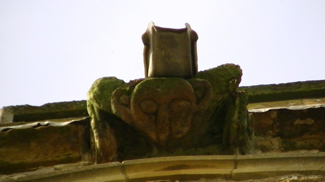 a gargoyle with a lead water spout on its back looking down from a church tower.
gargoyle is crouched with a crudely carved face, big ears.