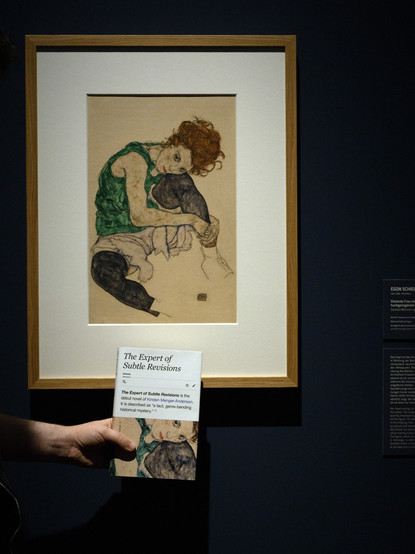 Ein Gemälde in einer Ausstellung, in Vordergrund hält eine Hand ein Buch mit dem Titel "The Expert of Subtle Revisions". Das Titelbild des Buches ist ein Ausschnitt aus dem Gemälde. Beim Gemälde handelt es sich um ein Bild von Egon Schiele. 