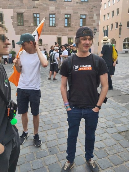 Menschen auf einem Platz, manche mit einer Fahne. Im Vordergrund ein Mann mit einem T-Shirt der Piratenpartei.