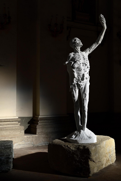 A statue of a dessicated mostly skeletal human, standing upright. He holds his left hand aloft, and stares up at his hand which contains an indeterminate object (from this angle, in which his body mostly faces the camera) but IMO may represent his heart.