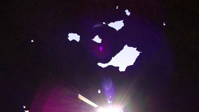 rust holes from standing inside a giant thin steel cylinder looks a bit like a face. sunlight shining through a lower hole causing beautiful purple lenz flare.