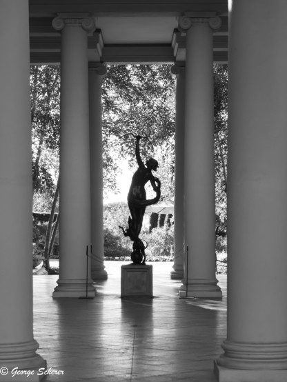 This is a black and white photo of a statue seen in silhouette, flanked by large columns.  The statue is a representation of the Roman goddess Diana, holding a bow, with a hunting dog at her feet.