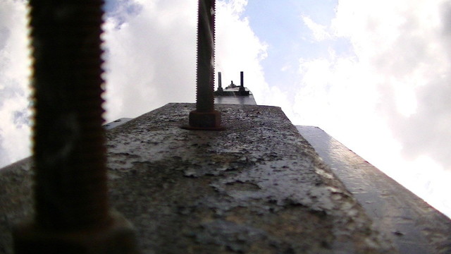 looking upwards at one arm of an open draw bridge from the counter weight. it is wooden with thick steel plates and long threaded bolts sticking out. the whole lot is painted black the sky is cloudy with patchy sun. a slight ghost of a spiders web is barely visible in the partial sunlight. my "G" key is still intermittent pmo