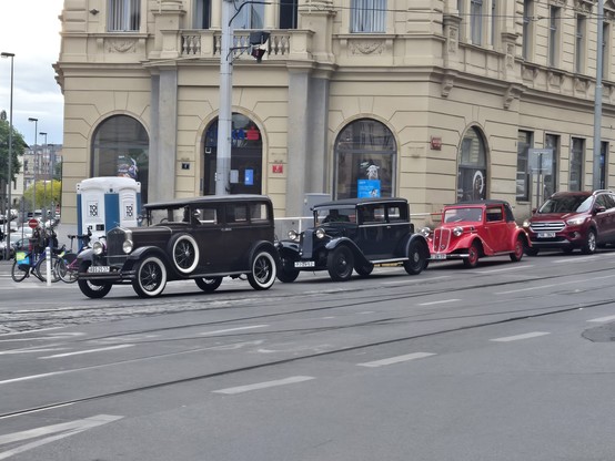 3 cars build around 1920 on street, behind them, there's a Ford from current era.

First one is black, have spare on doors and white tires.
Second one is also black with nice curved fender on the side.
Third one is red with side mirrors on fenders.