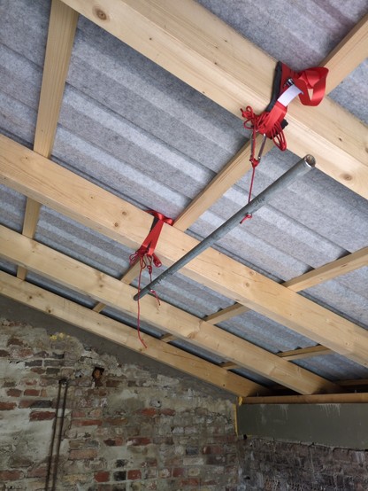Hängende Klimmzugstange mit rotem Paracord und roten Spanngurten an hellen Dachbalken unter einem Blechdach