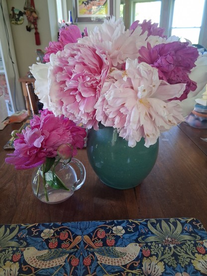 A vase and small jar full of peonies. They range in color from light pink to almost magenta. Ridiculous flowers are so heavy they bend their stems right to the ground.